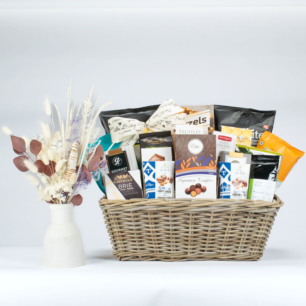 Rectangular ratan gift basket with chocolates, chips cookies and crackers. Neutral dried flower bouquet in wooden vase.
