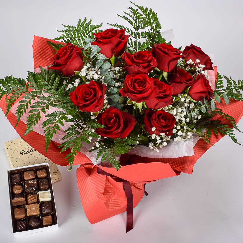 A Dozen Red Roses Wrapped In Red Textured Floral Paper With A Box Of Assorted Chocolates